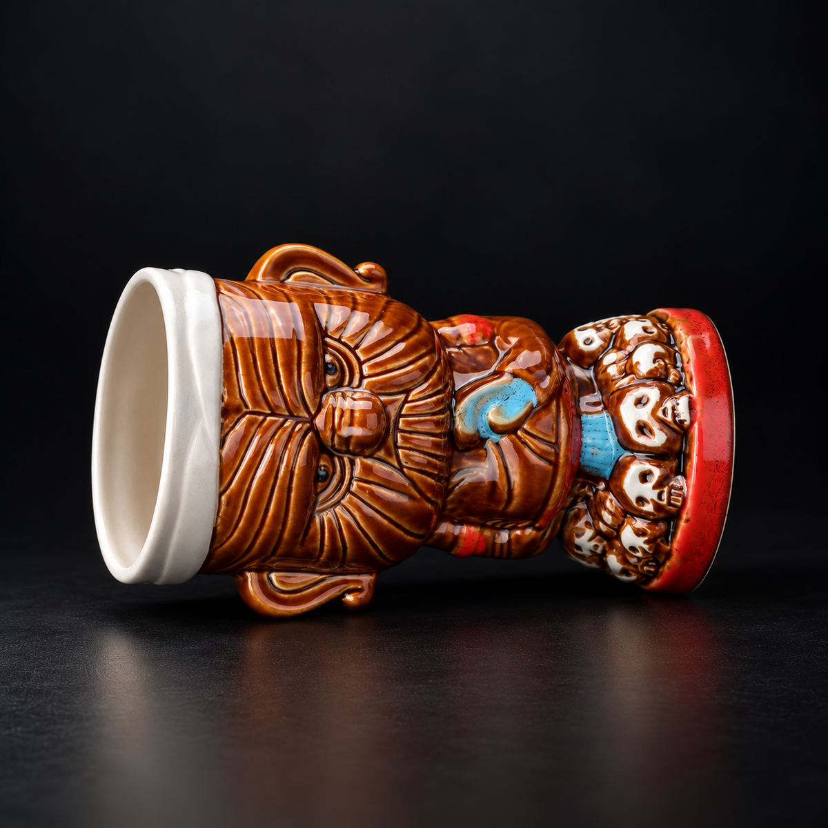 A decorative ceramic mug shaped like a stylized face with carved facial features, a white interior, and colorful blue, red, and brown painted details on a dark reflective surface.