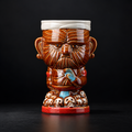 A ceramic mug shaped like a stylized warrior head with a white bandage on the head, brown carved facial features, a red base, and small skull details, photographed against a dark studio background.