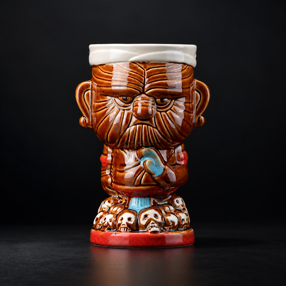 A ceramic mug shaped like a stylized warrior head with a white bandage on the head, brown carved facial features, a red base, and small skull details, photographed against a dark studio background.