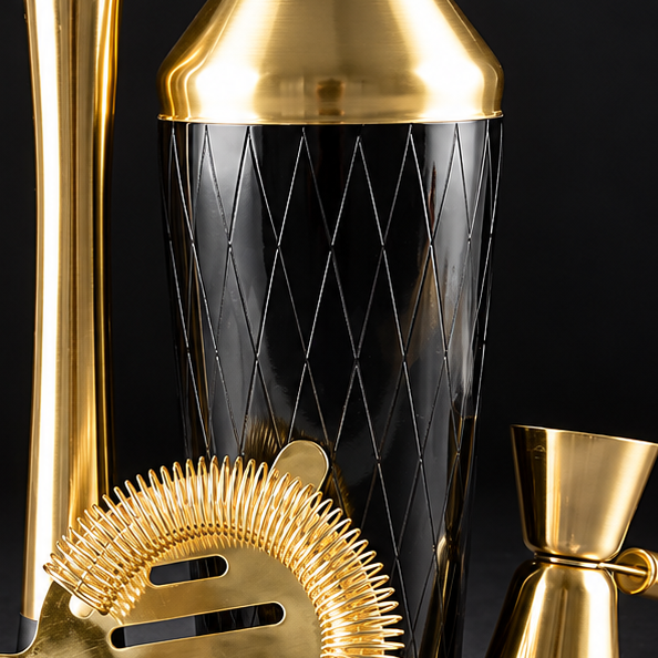 A gold and black cocktail mixing set arranged on a dark surface against a black background, including a shaker, strainer, jigger, spoon, and muddler.