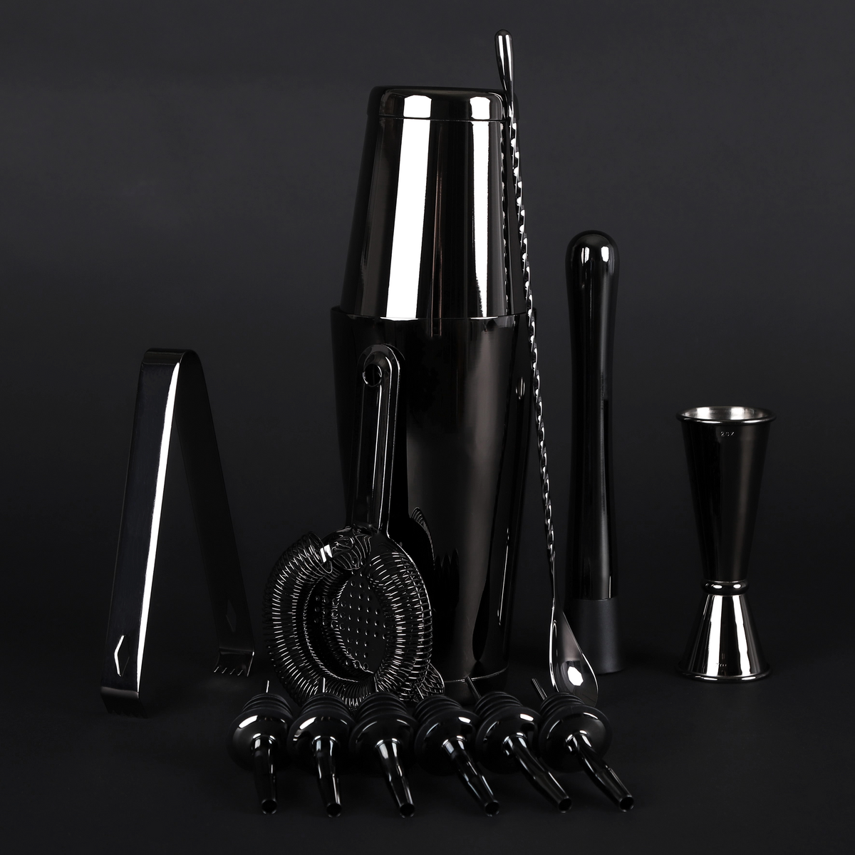 A professional bar tool set in polished black and silver stainless steel, including a shaker, muddler, jigger, strainer, and pourers, arranged on a dark studio background.